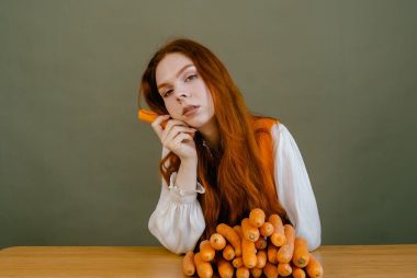 Portrait of a red-haired woman holding carrots, sitting at a table against a green background.